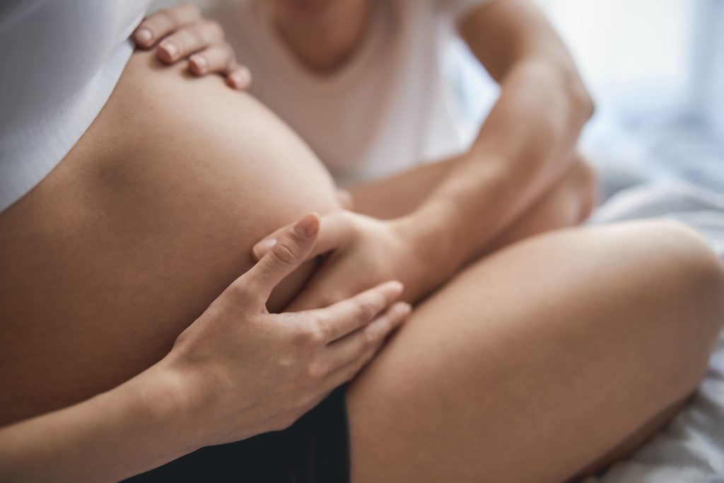 Nahaufnahme des Bauches einer schwangeren Frau, andere Frau fasst ihr an den Bauch